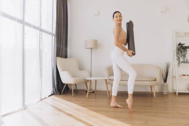 Elinde yoga minderi tutan genç bir kadın, rahat bir oturma odasında egzersiz yapmaya hazırlanıyor, sağlıklı bir yaşam tarzı ve form rutinini somutlaştırıyor.