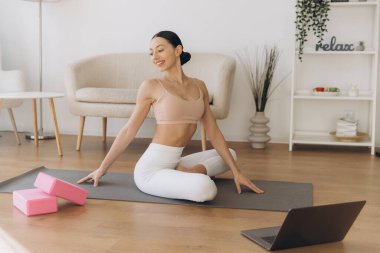 Yoga yapan sağlıklı bir kadın dizüstü bilgisayarındaki dersleri takip ederek evde sakin bir egzersiz ortamı yaratıyor.