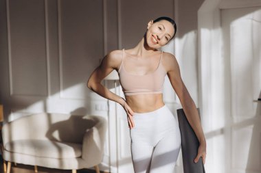 Spor kıyafetleri giyen, yoga minderi tutan ve güneşli bir odada egzersiz yaptıktan sonra gülümseyen bir kadın.