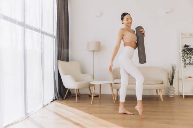 Elinde yoga minderi tutan sağlıklı bir kadın, sıcak oturma odasında canlandırıcı bir egzersiz için hazırlanıyor, sağlıklı bir yaşam tarzını kucaklıyor.