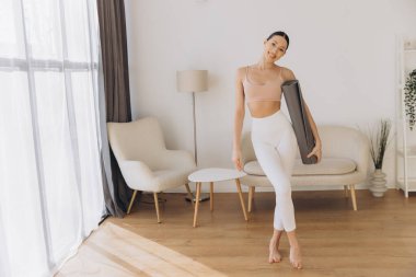 Elinde yoga minderi tutan sağlıklı bir kadın ev antrenmanına hazırlanıyor sağlığını ve sağlıklı yaşam tarzını destekliyor.