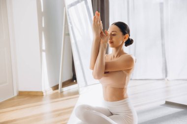 Garudasana 'da ya da Kartal Kolları pozunda yoga yapan bir kadın, parlak bir odada ayna ile oturuyor.