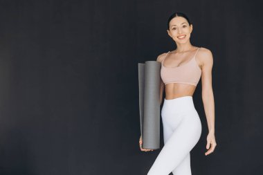 Gülümseyen yoga hocası yoga minderi tutarken sınıfa hazırlanırken dikkat çekici bir siyah arkaplan, kendine güven ve pozitiflik yayıyor.