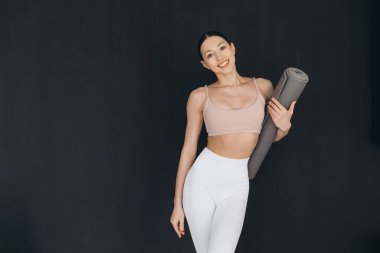 Siyah arkaplana karşı yoga minderi tutarken gülümseyen sportif kadın, canlandırıcı bir yoga pratiği ve dikkatli bir egzersiz için hazırlandı.