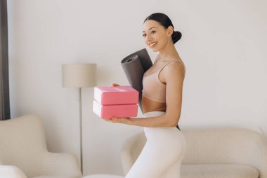 Yoga hocası, elinde yoga blokları ve minder tutarken gülümsüyor, doğal ışıkla dolu sıcak bir oturma odasında huzurlu bir anın tadını çıkarıyor.