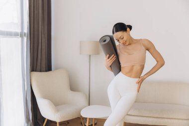 Elinde yoga minderi tutan sağlıklı bir kadın, evinde rahat oturma odasında canlandırıcı bir antrenmana hazırlanıyor, sağlıklı bir yaşam tarzını kucaklıyor.