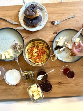 Türk kahvaltı masası. İnsanların elleri hamur işlerini, sebzeleri, yeşillikleri, zeytinleri, peynirleri, kızarmış yumurtaları, baharatları, reçelleri, balı, bakır demlikte çayı ve lale bardaklarını, geniş kompozisyonu alıyor.