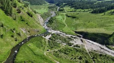 Yeşil bir vadi, ağaçlar ve bir kamp alanında park edilmiş bir araçla birleşen iki dar dağ nehrinin insansız hava aracı görüntüsü. Doğu Kazakistan