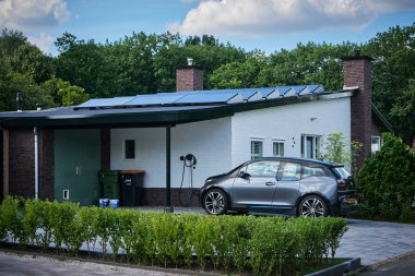 Güneş panelleriyle donatılmış bir evde şarj aleti olan elektrikli bir araç. Çevre dostu bir ortamda sürdürülebilir yaşam ve yenilenebilir enerji çözümlerinin modern bir örneği.