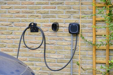 Hollanda: sürdürülebilir enerji çözümlerini ve elektrik araç altyapısının artan eğilimini sembolize eden, bahçe bitkileriyle dolu sarı tuğla bir duvara monte edilmiş bir EV şarj kutusu, ince zeka kutusu.