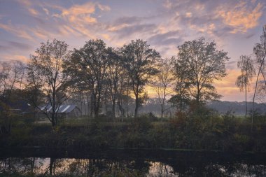 Tranquil landscape in Drenthe featuring trees silhouetted against a colorful sunset sky, with reflections on calm water, capturing the beauty of nature. clipart