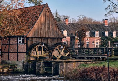 A beautifully preserved historic watermill in Lage, Germany, featuring wooden wheels and traditional timber and brick construction near a flowing river. clipart