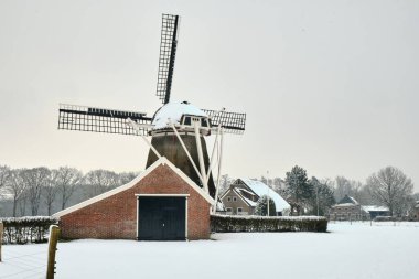 Oudemolen, Drenthe, Netherlands - February 12, 2025. Traditional Dutch windmill De Zwaluw seen over her red brick shed, covered in snow on a serene winter day, surrounded by white fields. clipart