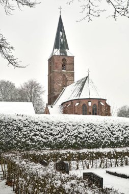 A historic brick church with a tall steeple in Rolde, Netherlands, surrounded by a snow-covered cemetery and bare winter trees on an overcast day. clipart