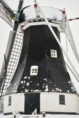 A historic windmill in Rolde, Netherlands, covered in fresh snow, surrounded by winter trees and a peaceful rural setting. clipart