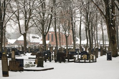 A peaceful cemetery in Rolde, Netherlands, covered in fresh snow, with an old church and bare trees in the background on a quiet winter day. clipart