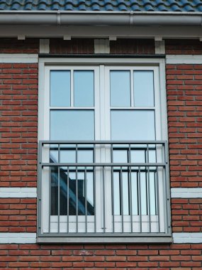 Beyaz çerçeveli bir penceresi ve kırmızı tuğlalı ön cephesinde metal parmaklıkları olan modern bir Fransız balkonu çevresindeki binaları ve gökyüzünü yansıtıyor..