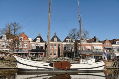 6 Mart 2025 - Dokkum-Hollanda: Dokkum kanalı boyunca demirlemiş iki direkli beyaz bir koftjalk gemisi, tarihi Hollanda konakları ve liman terası.