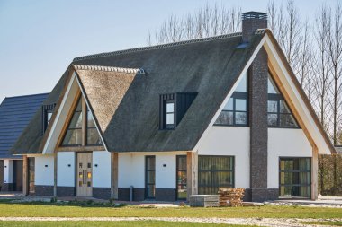 1 Nisan 2025 - Nieuweroord-Hollanda: Geniş bir arazide sazdan bir çatısı olan yeni inşa edilmiş modern villa, inşaat malzemeleri hala görünür, henüz tamamlandı.