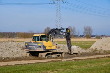 5 Mart 2025 - Warga-Hollanda: Volvo sürüngen kazıcısı, geniş toprak tepecikleri, geçici çelik plaka yolu ve arka planda Frizya manzarası boyunca uzanan elektrik hatları ile devam eden polder çalışmaları.