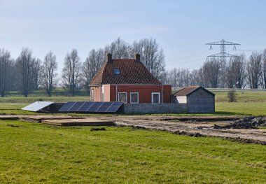 5 Mart 2025 - Warga-Hollanda: Modern uzantılı yel değirmeni yakınlarındaki küçük kırsal ev, çit olarak zemin monteli güneş panelleri ve polder bakımı için geçici çelik yol.