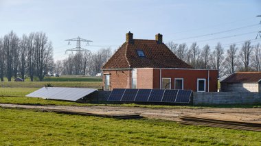 5 Mart 2025 - Warga-Hollanda: Modern uzantılı yel değirmeni yakınlarındaki küçük kırsal ev, çit olarak zemin monteli güneş panelleri ve polder bakımı için geçici çelik yol.