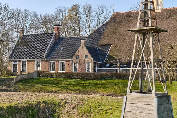 8 Mart 2025 - Ropta-Hollanda: Güneşli bir bahar gününde huzurlu bir Hollanda kırsalında, ön planda bir Amerikan yel değirmeninin çelik tabanına sahip tipik bir Frizya kop-hals-romp çiftlik evi.
