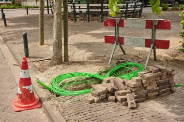 14 Mayıs 2025 - Oudelande-Hollanda: Yeşil kablolar, istiflenmiş tuğlalar ve kaldırım taşı bir sokakta hasarlı bir trafik konisi ile kırsal bir alanda fiber optik ağ kurulumu.