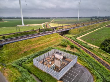 27 Mayıs 2025 - Moerdijk-Hollanda: Moerdijk demiryolu köprüsü yakınlarındaki bir tren istasyonunun havadan görüntüsü, yeşil alanlar ve bulutlu bir gökyüzü altında modern rüzgar türbinleriyle çevrili.