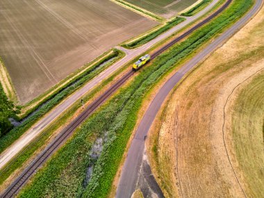 27 Mayıs 2025 - Moerdijk-Hollanda: Moerdijk demiryolu köprüsü yakınlarındaki yeşil alanlarda tek bir yük vagonunda seyahat eden sarı bir demiryolu lokomotifinin havadan görünüşü.