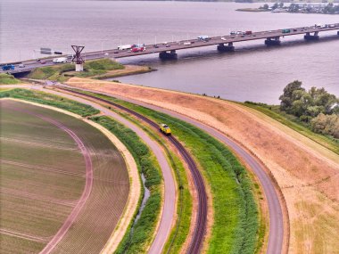 27 Mayıs 2025 - Moerdijk-Hollanda: Moerdijk Köprüsü 'nün havadan görünüşü ile sarı bir demiryolu lokomotifi yeşil alanlarda tek bir yük yolu üzerinde seyahat ediyor..