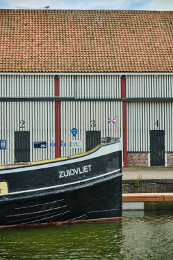 26 Mayıs 2025 - Zierikzee-Hollanda: Zierikzee limanındaki tarihi Stadstimmerwerf limanının önündeki rıhtım boyunca demirlemiş jeuzen bayrağıyla geleneksel siyah ve sarı bir römorkör yayı.