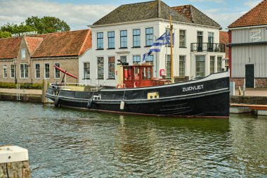 26 Mayıs 2025 - Zierikzee-Hollanda: Zierikzee limanındaki tarihi Stadstimmerwerf limanının önünde Zeeland bayrağı dalgalanan geleneksel bir siyah ve sarı römorkör.