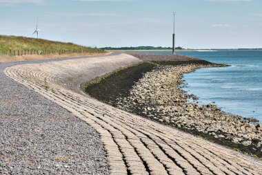 Hollanda, Zeeland 'deki Oosterschelde haliçleri boyunca uzanan eğri taşlı bir deniz yatağı, kıyı kayaları, berrak mavi su ve arka planda bir rüzgar türbini bulunuyor..