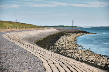 Hollanda, Zeeland 'deki Oosterschelde haliçleri boyunca uzanan eğri taşlı bir deniz yatağı, kıyı kayaları, berrak mavi su ve arka planda bir rüzgar türbini bulunuyor..