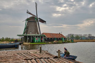 13 Nisan 2025 - Zaandam-Hollanda: Geleneksel bir Hollandalı değirmenci ve küçük bir teknedeki bir gönüllü, arka planda çalışan petrol değirmeni De Zoeker 'in tarihi kereste fabrikasında işlenmek üzere yüzen bir kütük seçtiler..