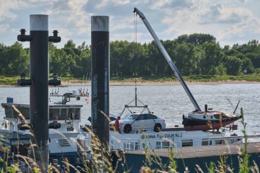 2 Haziran 2025 - Tolkamer-Hollanda: Bir iç kargo gemisi, dramatik yaz bulutları altında büyük demirleme direkleriyle Rhine otomobil rampasında bir araba boşaltıyor.