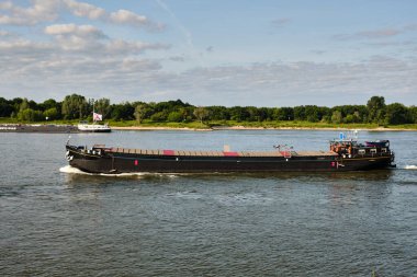 3 Haziran 2025 - Tolkamer-Hollanda: Küçük bir vintage kargo mavnası Rhine Nehri 'ne açılır, halen modern nakliyede aktiftir, arka planda başka bir gemi ve yeşil kıyı şeridi vardır..