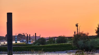 Gün batımında Tolkamer yakınlarında huzurlu bir nehir manzarası. Kargo gemileri, yeşil bankalar, çizgili bir deniz feneri ve yumuşak turuncu gökyüzü ile sakin bir atmosfer yaratıyor..