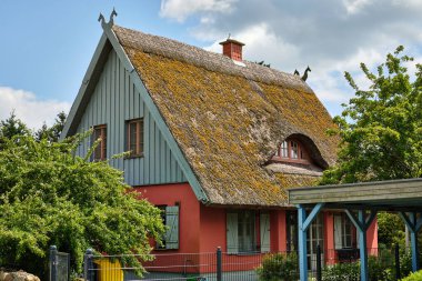 16 Haziran 2025 - Kamminke-Almanya: Kuzey Alman tarzında, yosun kaplı çatısı ve renkli cepheli, parçalı bulutlu bir gökyüzü altında yeşilliklerle çevrili geleneksel sazdan tatil evi.