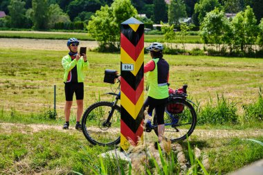 16 Haziran 2025 - Kamminke-Almanya: Kadın, Swinemunde yakınlarındaki Polonya sınırına işaret eden Alman bayrağının yanında poz verirken bisiklet kıyafeti giymiş bir çift fotoğraf çekiyor.