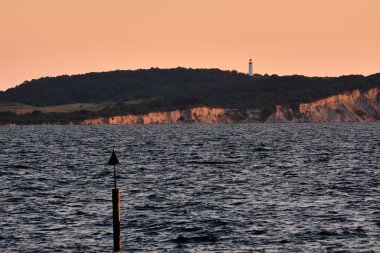Almanya 'nın Rugen kenti yakınlarındaki Hiddensee adasındaki dik tebeşir kayalıkları ve deniz fenerinin önündeki seyrüsefer fenerinin akşam görüşü, sakin bir yaz günbatımı sırasında sakin Baltık Denizi üzerinde turuncu gökyüzü.