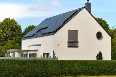 15 Temmuz 2025 - Klockenhagen-Almanya: Çatı katı güneş panelleri, yuvarlak pencereler, kapalı Fransız balkonu ve parçalı bulutlu bir gökyüzü altında düzgünce budanmış bir çit.