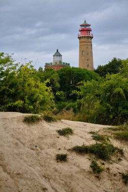 9 Temmuz 2025 - Putgarten-Almanya: Rugen adasında bulutlu bir gökyüzünün altında yeşil ağaçlar ve çalılarla çevrili iki tarihi tuğla deniz feneri.