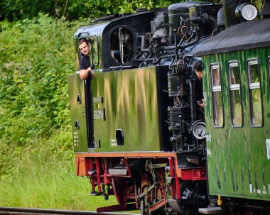 6 Temmuz 2025 - 99. sınıf buhar lokomotifi Rasender Roland Miras demiryolunun genç bir mühendis taksiden dışarı bakarken ve buhar kaçarken orman eğrisi boyunca ilerlemesi.