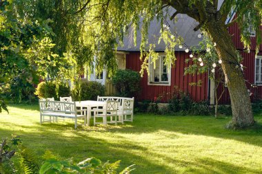 İsveç kır evi bahçesi beyaz masa, sandalyeler, yapraklı ağaç, açık hava lambası ve yaz güneşinde arka planda giriş..