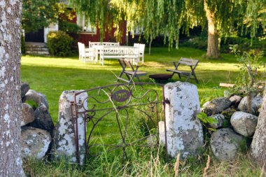 İsveç kır evi bahçesi, beyaz masa, sandalyeler, yapraklı ağaç, şömine ve yaz güneşi girişi..