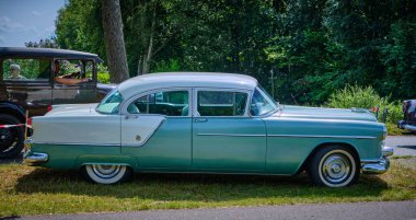 17 Ağustos 2024 - Ruinerwold-Hollanda: Eski zamanların geçit töreninde çimenli bir yol kenarına park edilmiş eski model Oldsmobile 88, arka planda diğer klasik arabaların geçişi ile birlikte.