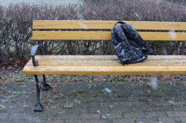 Snow falls gently on a yellow bench in a serene park, where a black backpack rests unattended, evoking a peaceful winter atmosphere. clipart