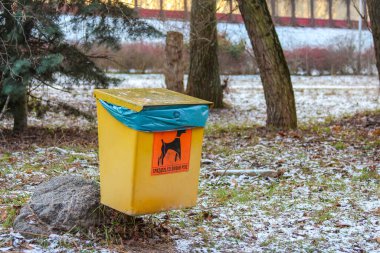 Parlak sarı bir köpek atığı, güvenilir hayvan sahiplerinin kullanması için sessiz bir parkta, kar ve ağaçların arasında bir kayanın üzerinde duruyor..